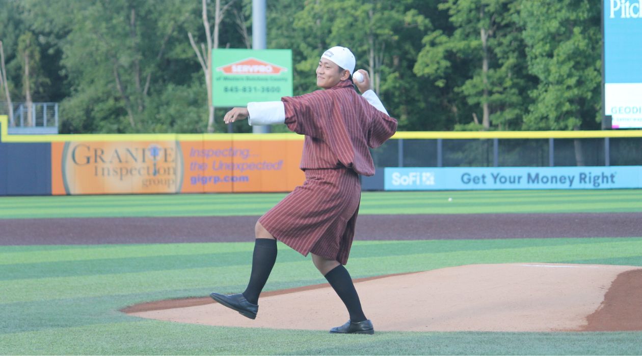 A young ballplayer from the Kingdom of Bhutan, wearing traditional Bhutanese clothes and a baseball cap on backwards, throws out a ceremonial first pitch prior to the Hudson Valley Renegades game at Heritage Financial Park in Wappingers Falls on Bhutan Night 2024